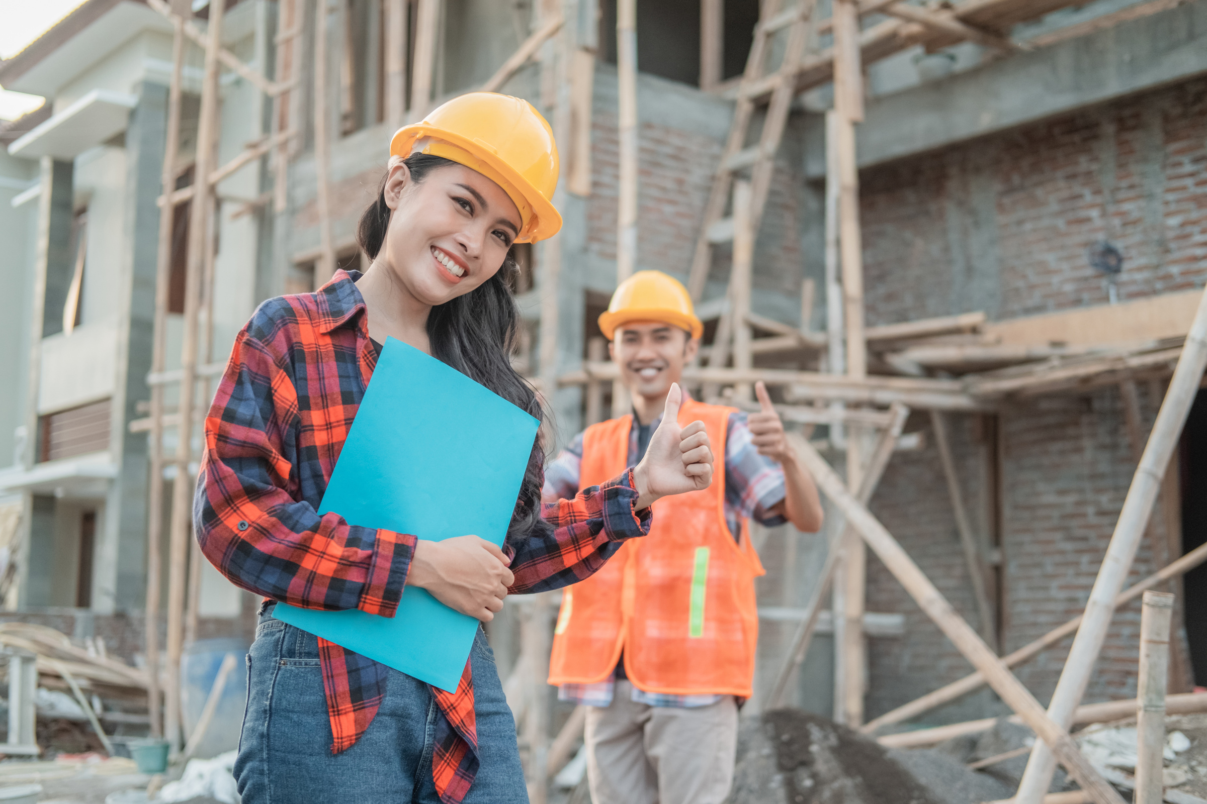 Asian Construction Worker Smiling at the Camera Wearing a Safety Helmet with a Thumbs up against the Background of a Male Contractor Standing